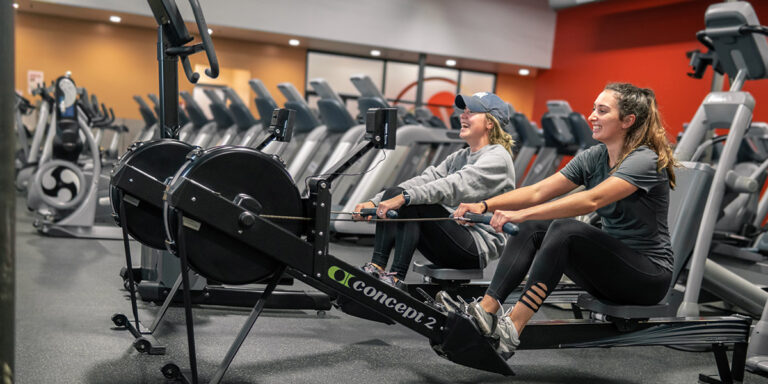 dos mujeres sonriendo juntas mientras utilizan las máquinas de ejercicios en el club de Conway